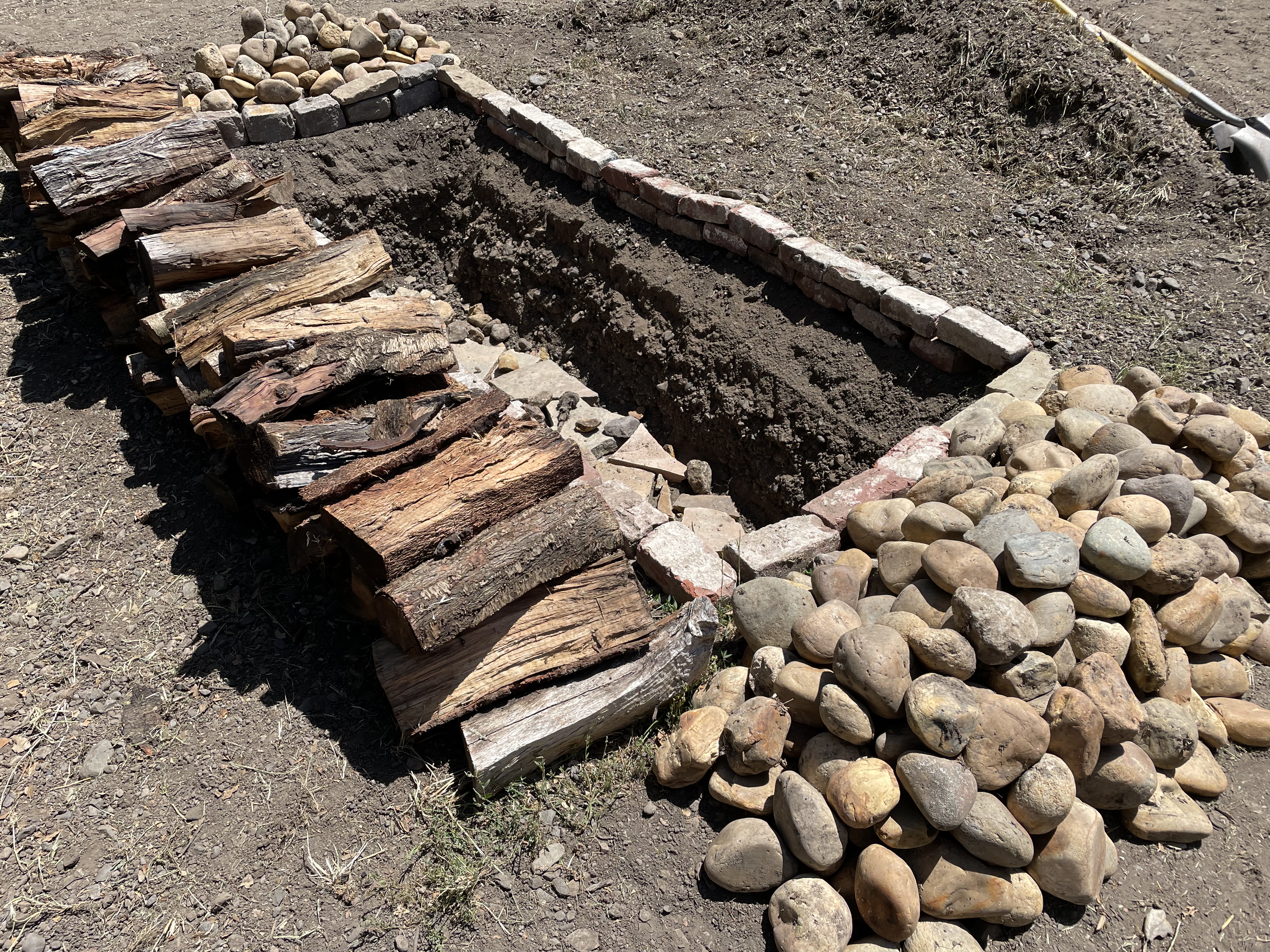 The hole, dug out by tractor and lined with bricks, fire wood on left and dirt to cover on right.